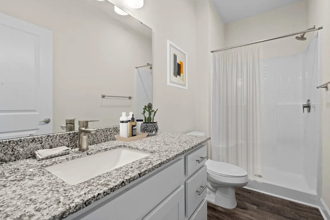 Modern bathroom with granite countertop sink, white cabinets, framed abstract art, toilet, and white tiled shower with curtain.