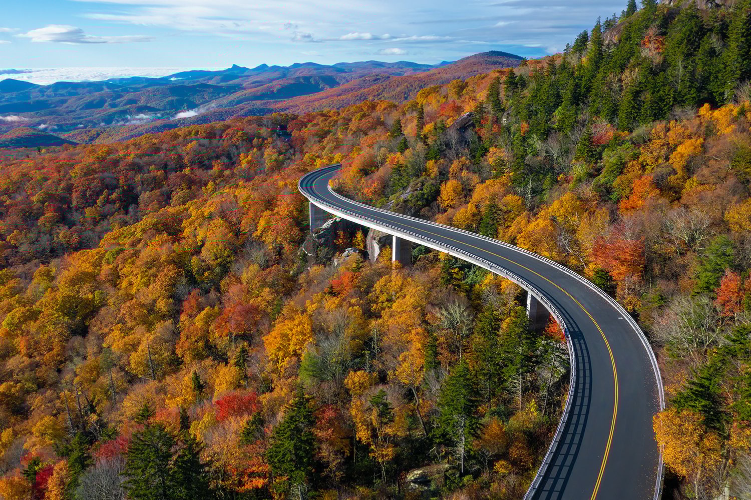 Curving mountain road bridge over vibrant autumn forest with colorful fall foliage and distant blue hills under a partly cloudy sky.