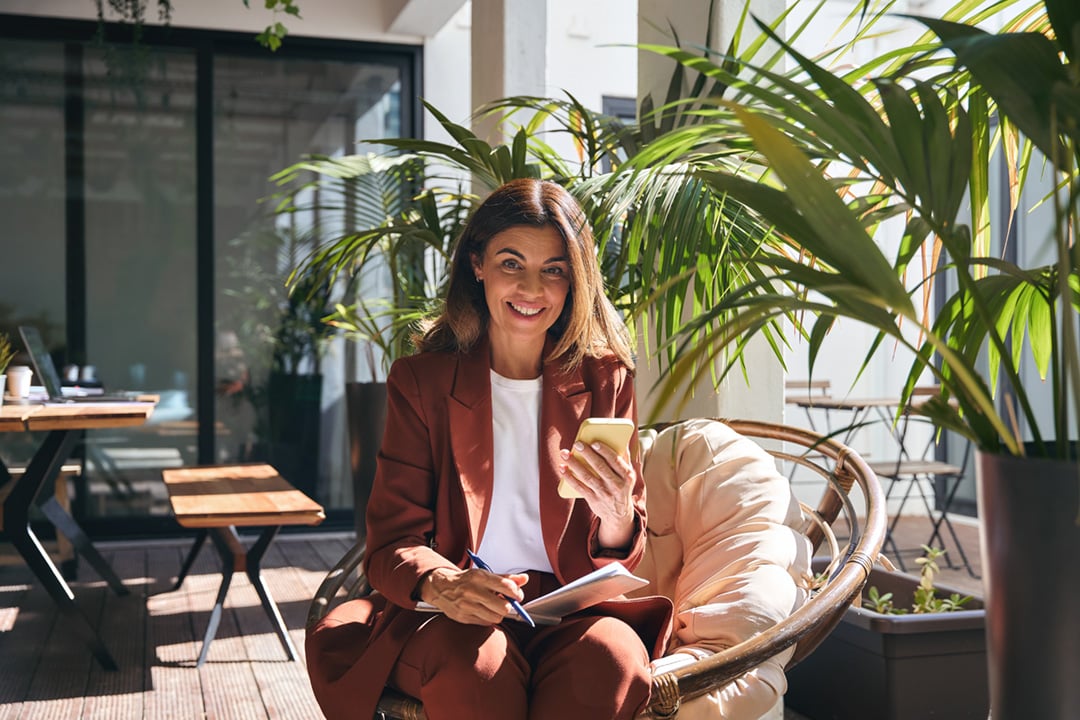 Smiling woman in a rust-colored suit sitting on a cushioned chair outdoors with plants, holding a smartphone and pen with notebook.