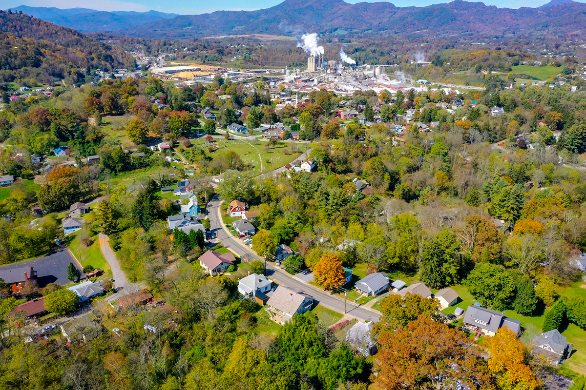 Aerial view of a small town with houses, trees, winding roads, and a factory emitting smoke, set against forested hills.