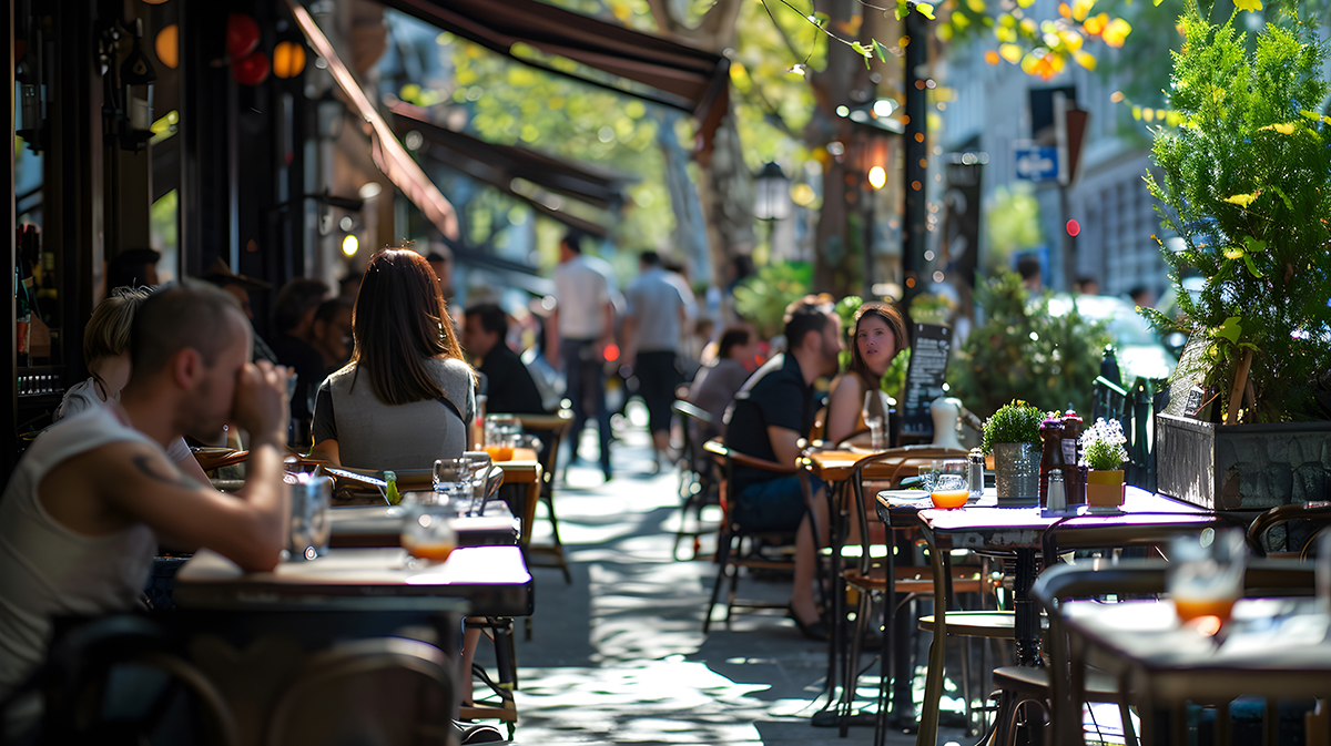 Outdoor café seating with people dining and walking on a sunlit urban sidewalk lined with trees and plants.