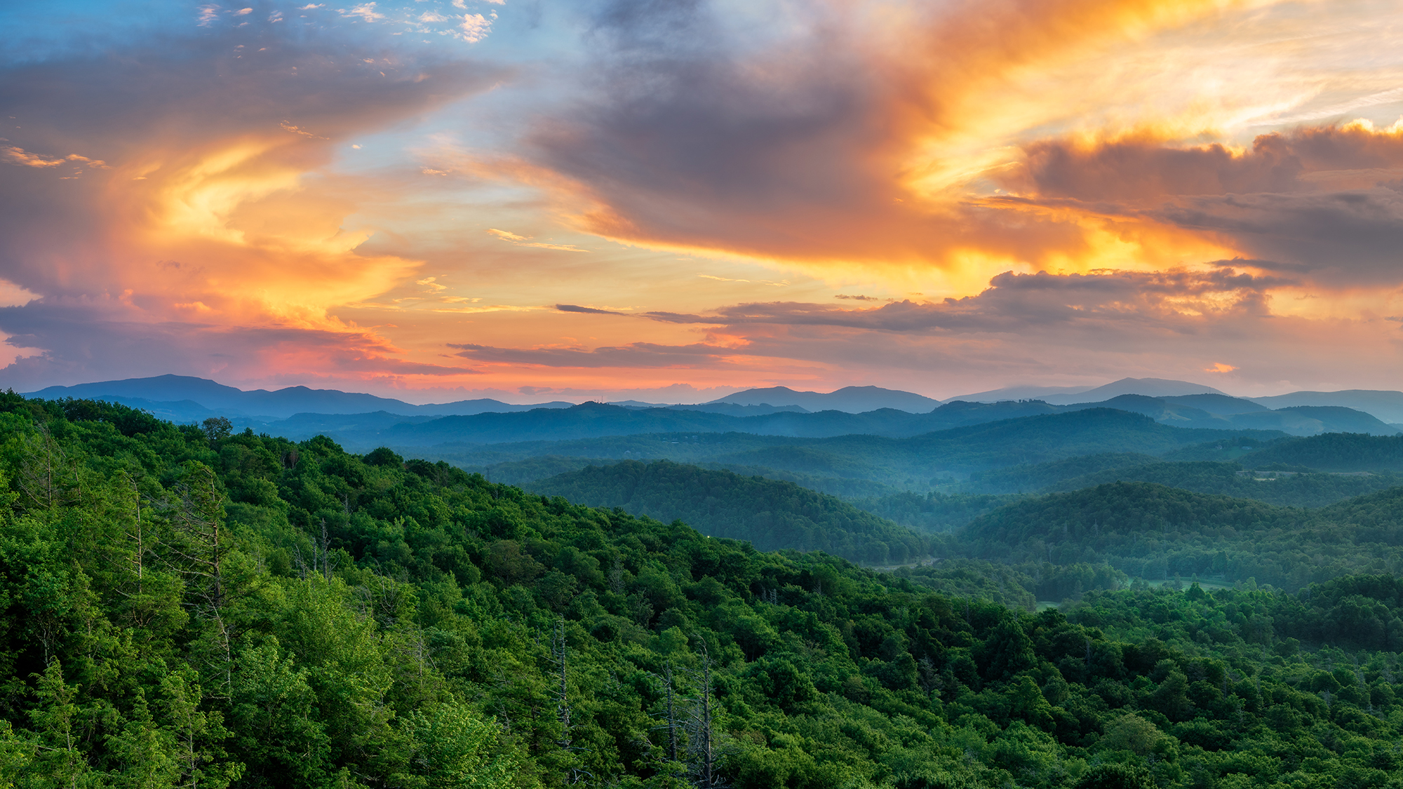 Sunset over layered blue ridge mountains with dense green forest in foreground and colorful cloudy sky.