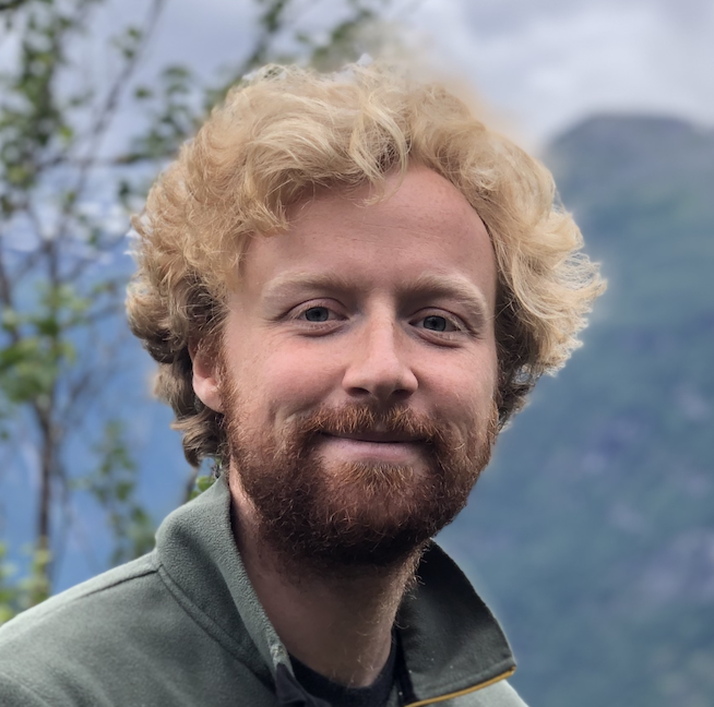 Aurélien Migeot. Smiling young man with curly blond hair and beard wearing a green jacket with blurred outdoor background.