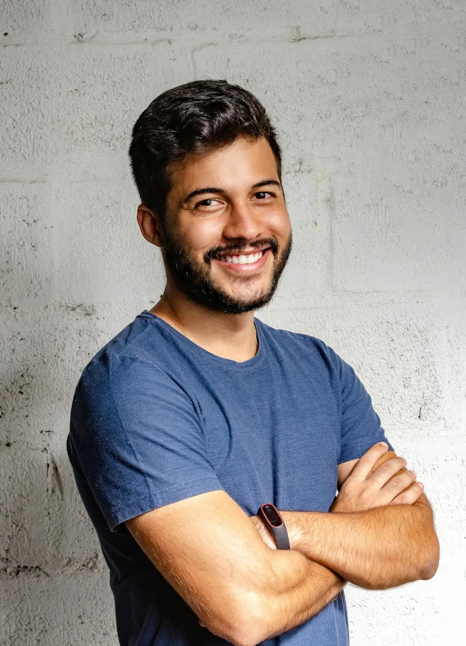 Smiling man with short dark hair and beard wearing a navy blue t-shirt, standing with arms crossed against a light textured wall.