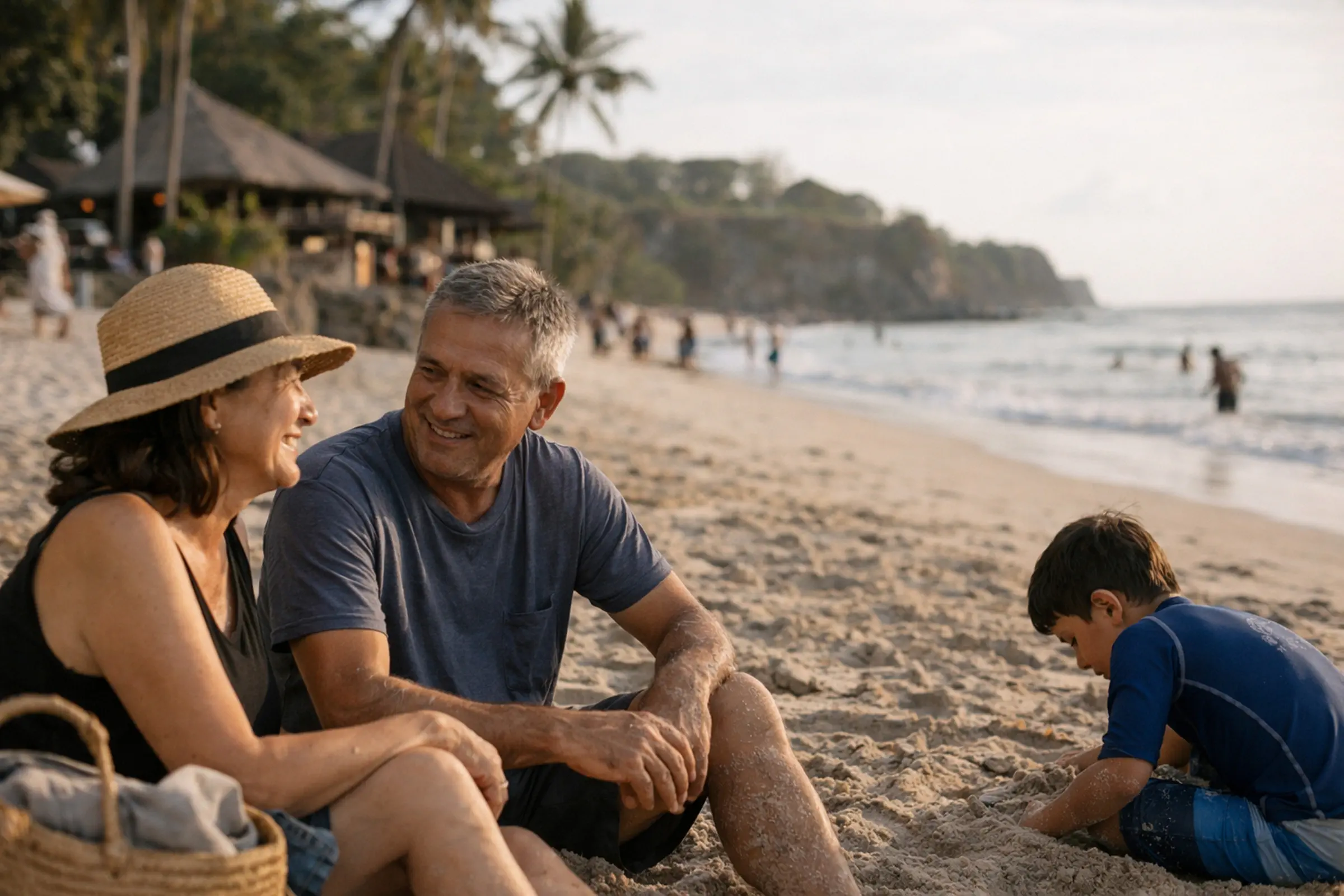 Middle-aged couple smiling and sitting on the sandy beach while a boy plays in the sand nearby during a sunny day.