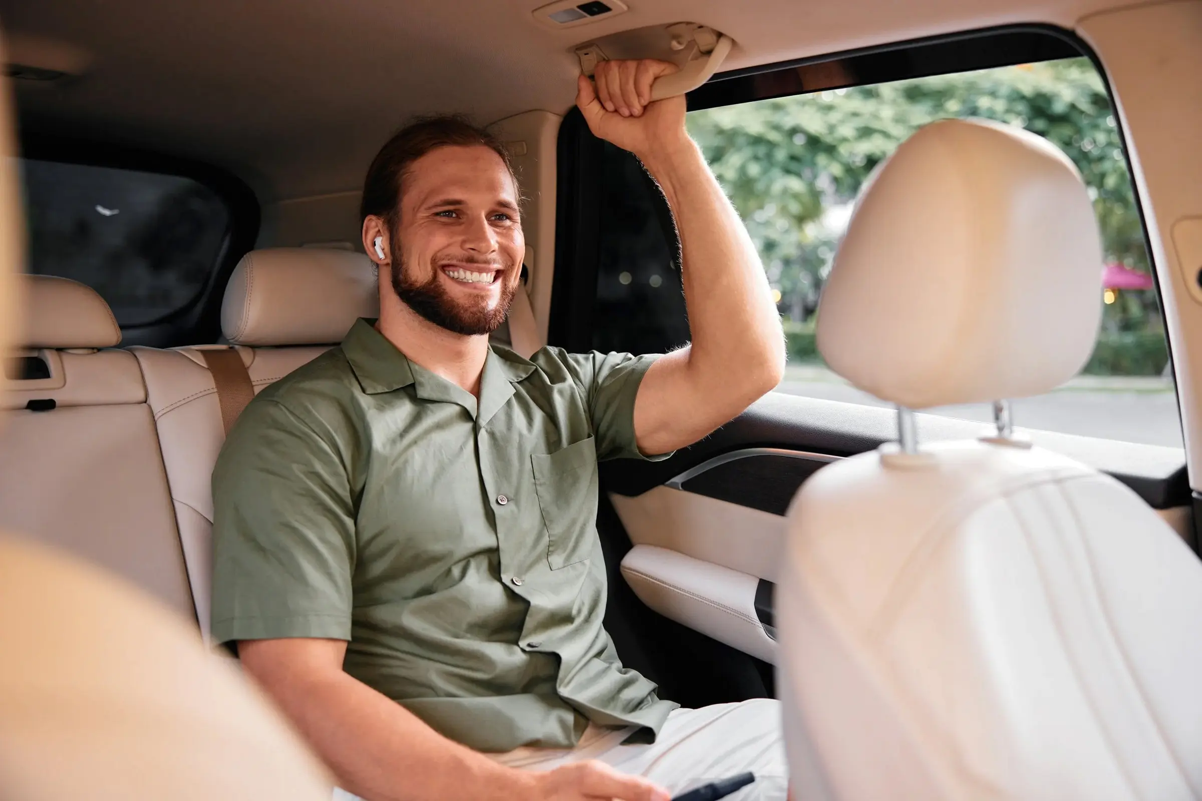 Smiling man wearing wireless earbuds, sitting in the back seat of a car holding the overhead handle.