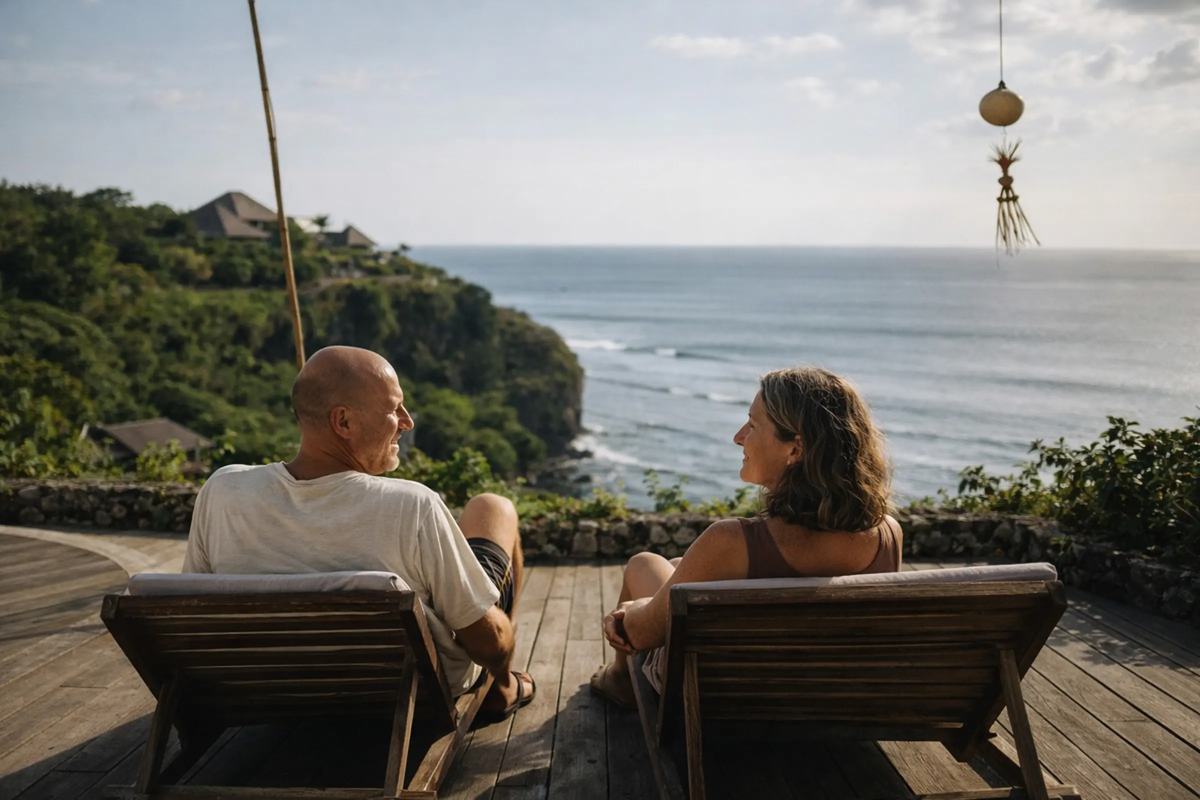 A man and woman sitting on wooden lounge chairs facing each other with a coastal cliff and ocean view in the background.