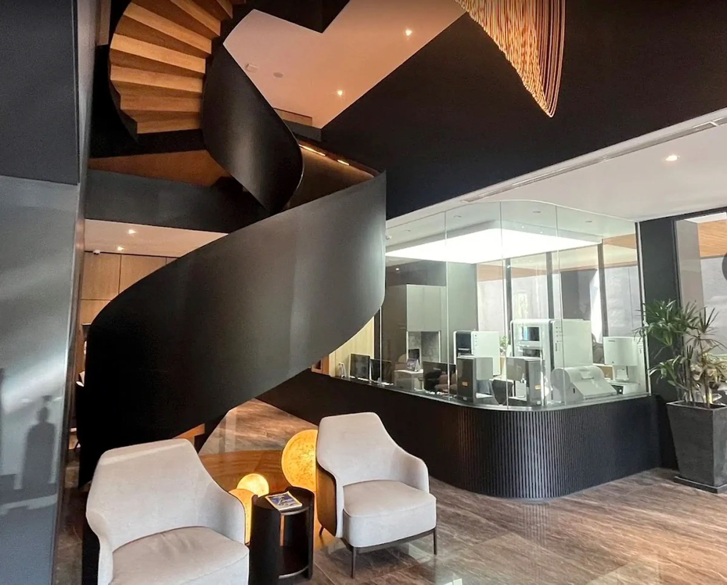 Modern interior with a black spiral staircase, two white armchairs, a small black side table, and a glass-walled office area.