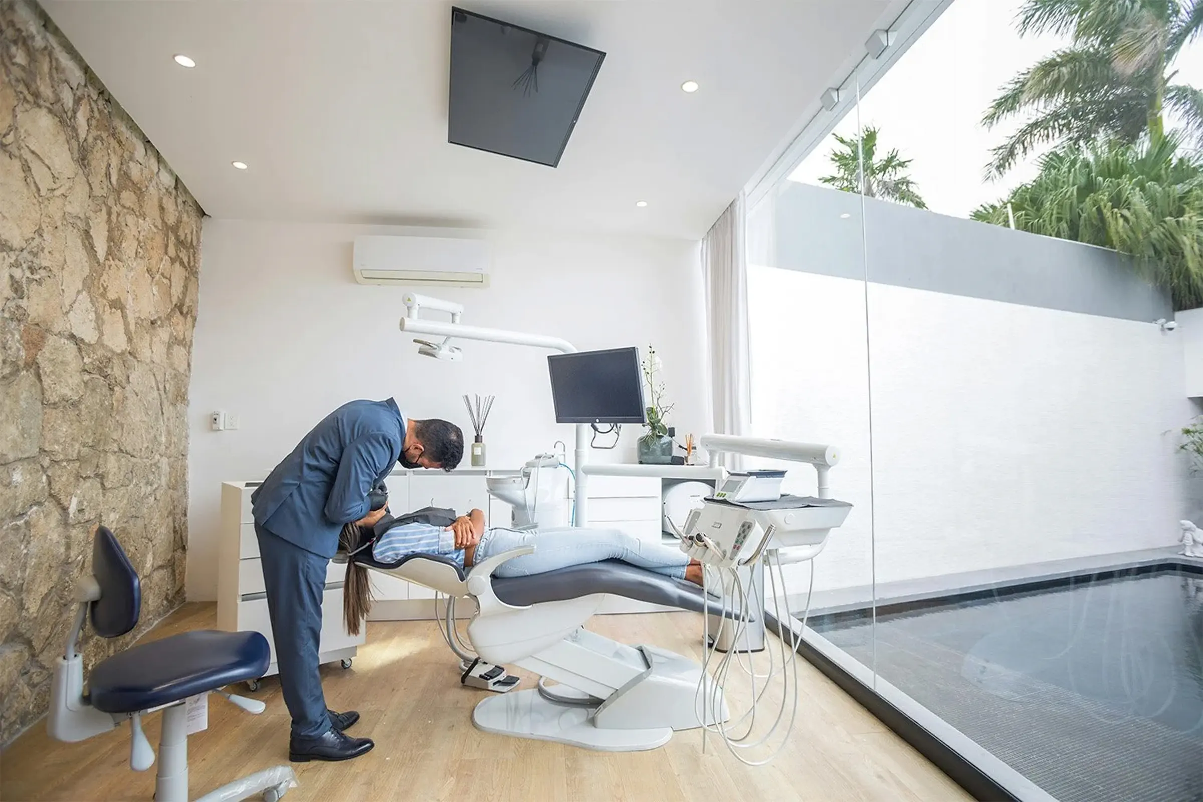 Male dentist in a suit examining a patient lying on a dental chair in a modern clinic with large window and stone wall.