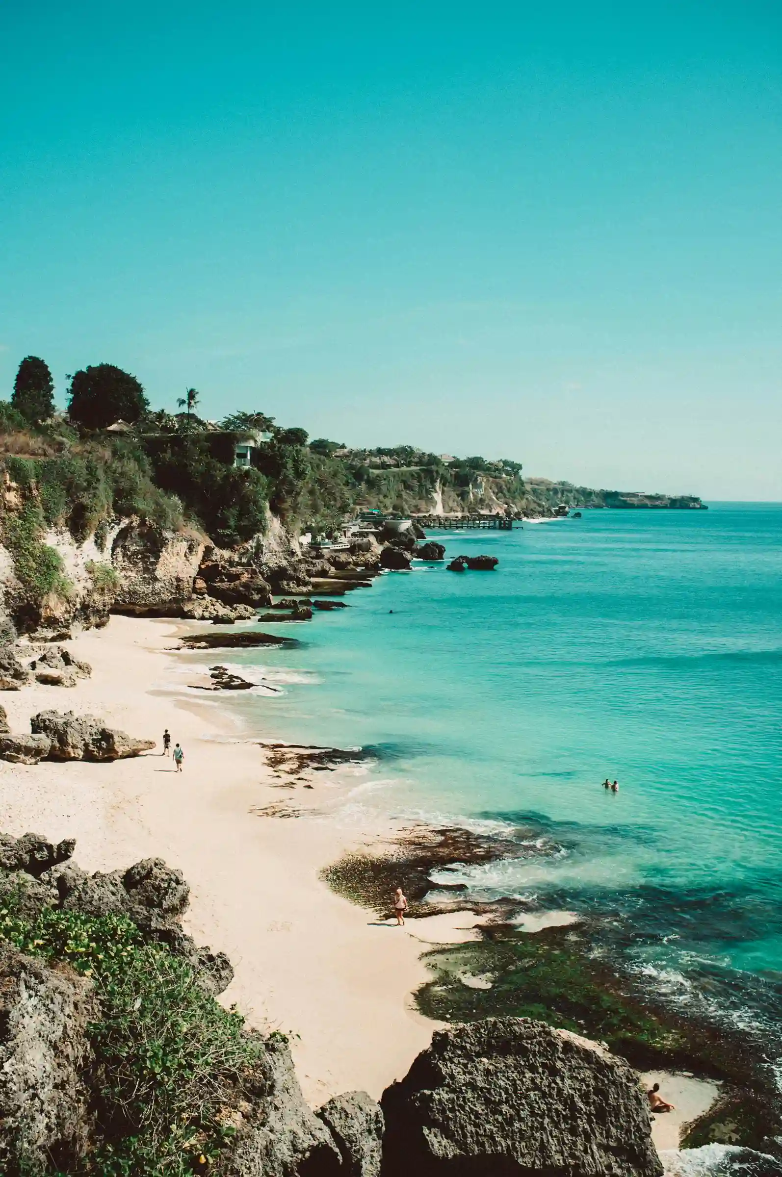 Rocky coastline with turquoise water, sandy beach, and a few people walking and swimming in a tropical setting.