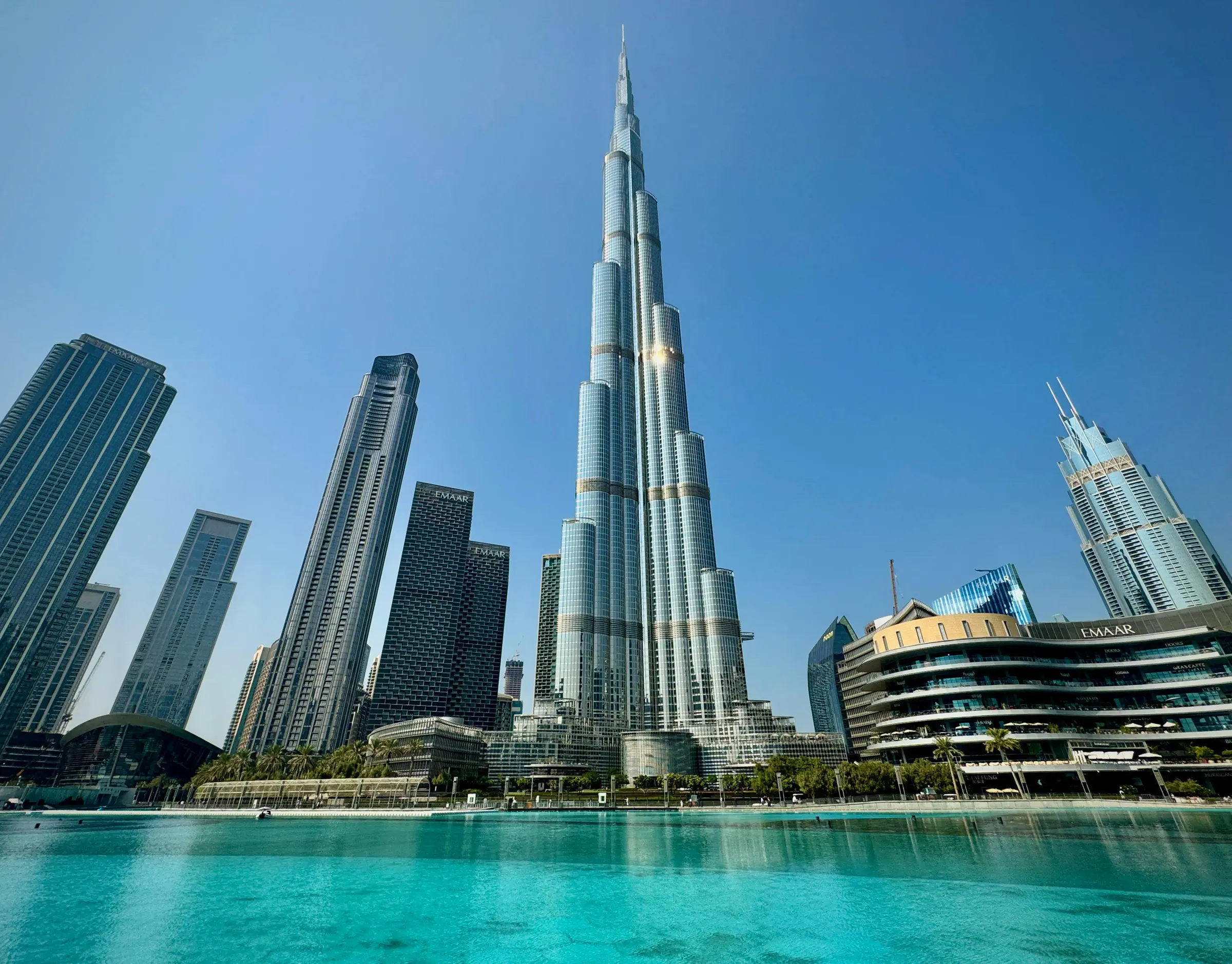 Burj Khalifa skyscraper towering over surrounding buildings with a turquoise water body in the foreground under a clear blue sky.