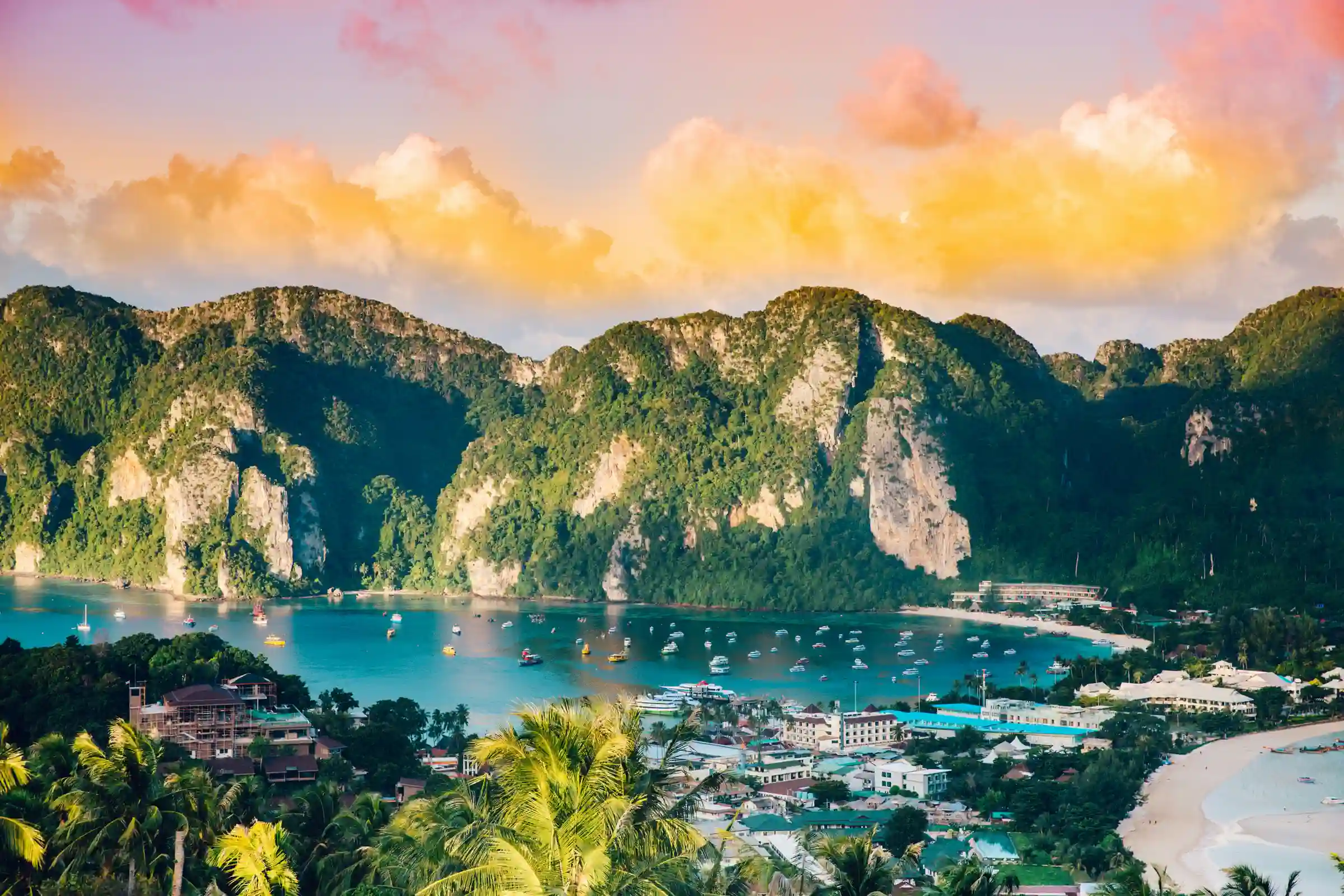 View of a tropical bay with turquoise water, numerous boats, lush green mountains, a sandy beach, and a coastal town under a colorful sunset sky.