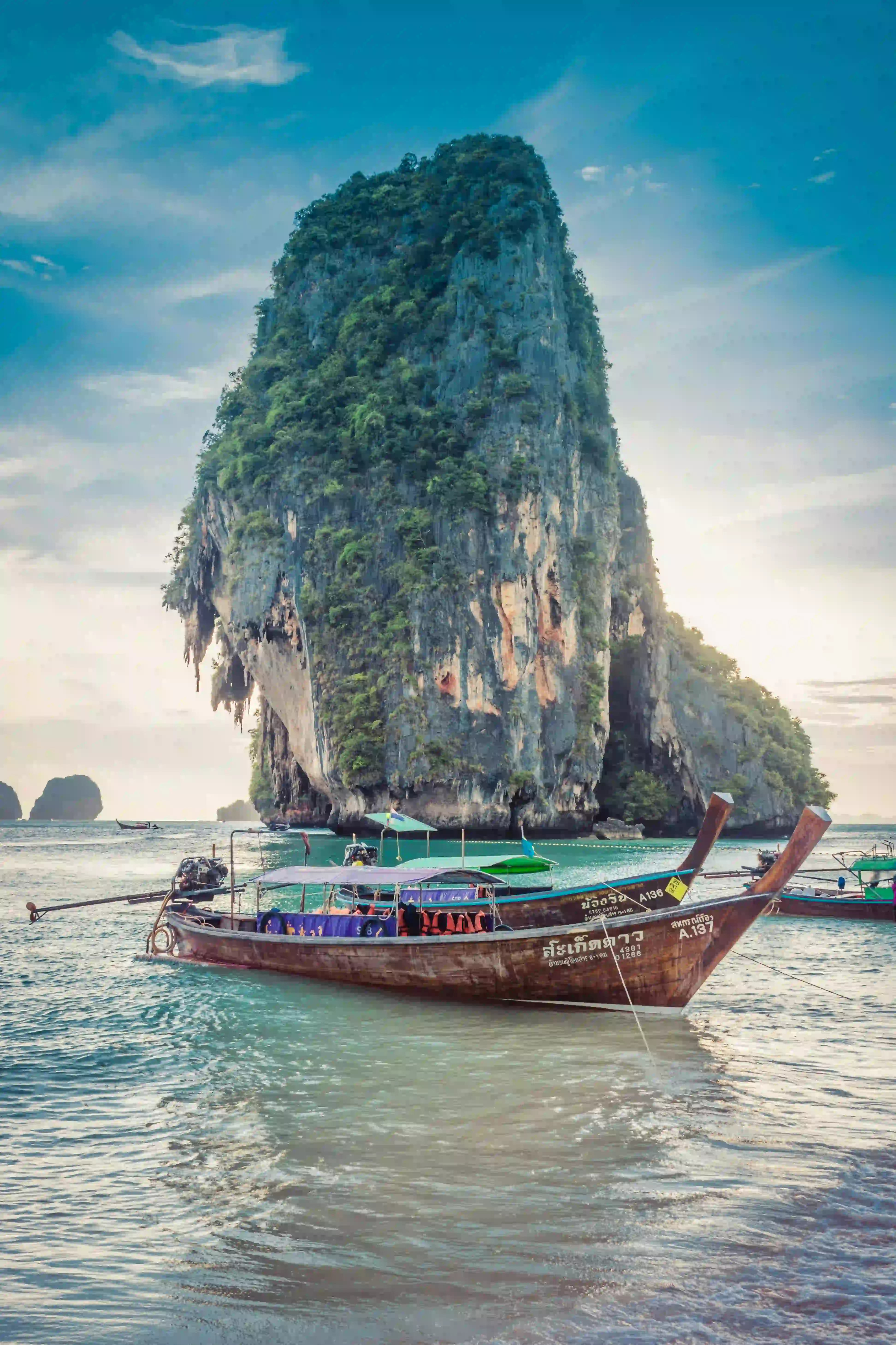 Traditional long-tail boat floating on turquoise water with a large limestone cliff covered in greenery in the background under a partly cloudy sky.