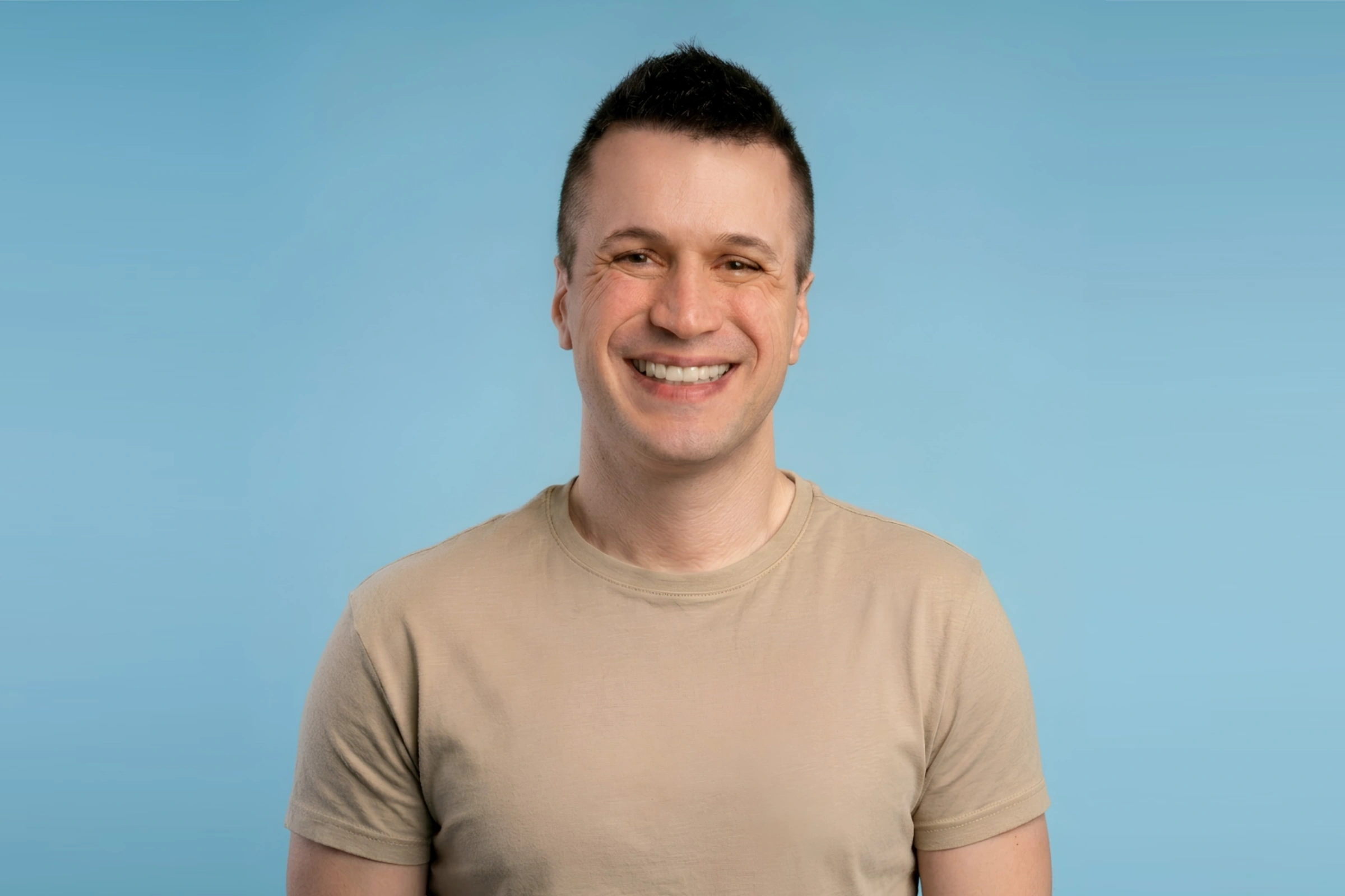 Smiling man with short dark hair wearing a beige t-shirt against a blue background.