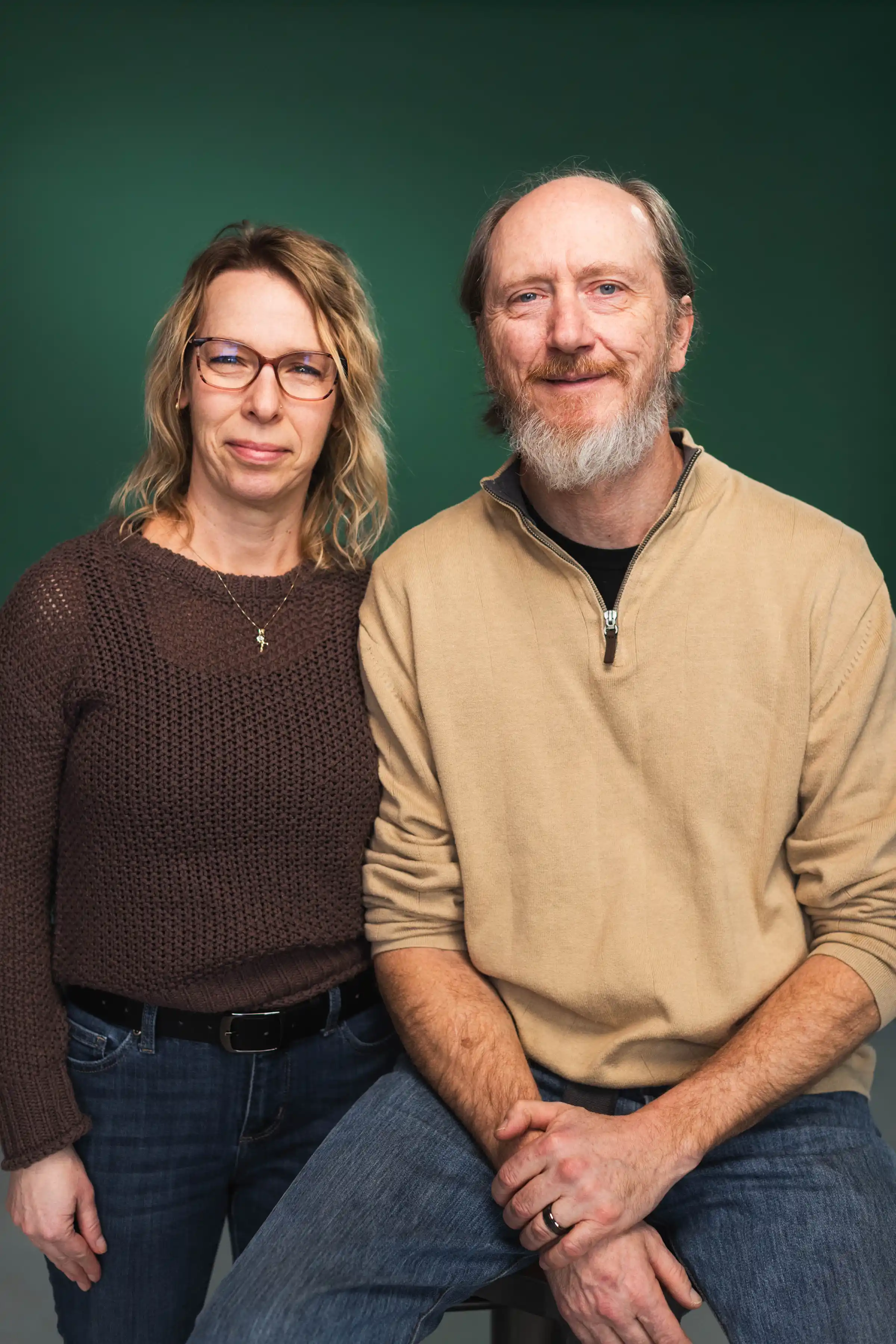 Smiling middle-aged man in a blue plaid shirt sitting on a stool with arm around a woman in glasses and a brown sweater standing beside him.