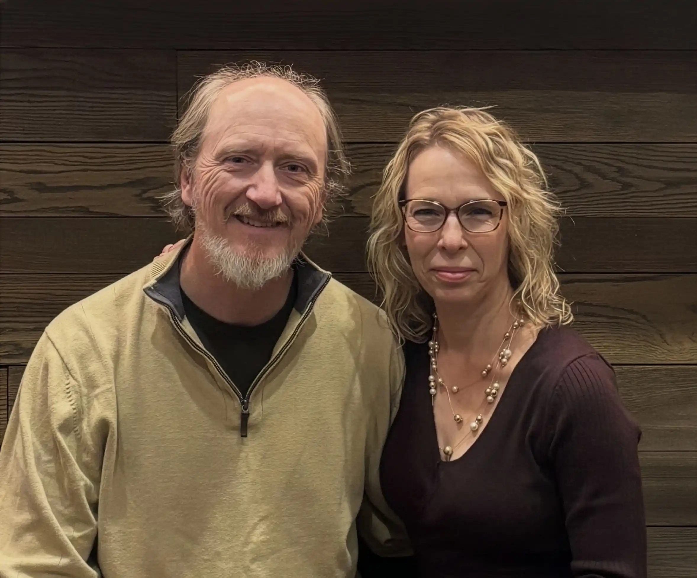 Smiling middle-aged couple posing in front of a green backdrop, with the man sitting and the woman standing beside him.