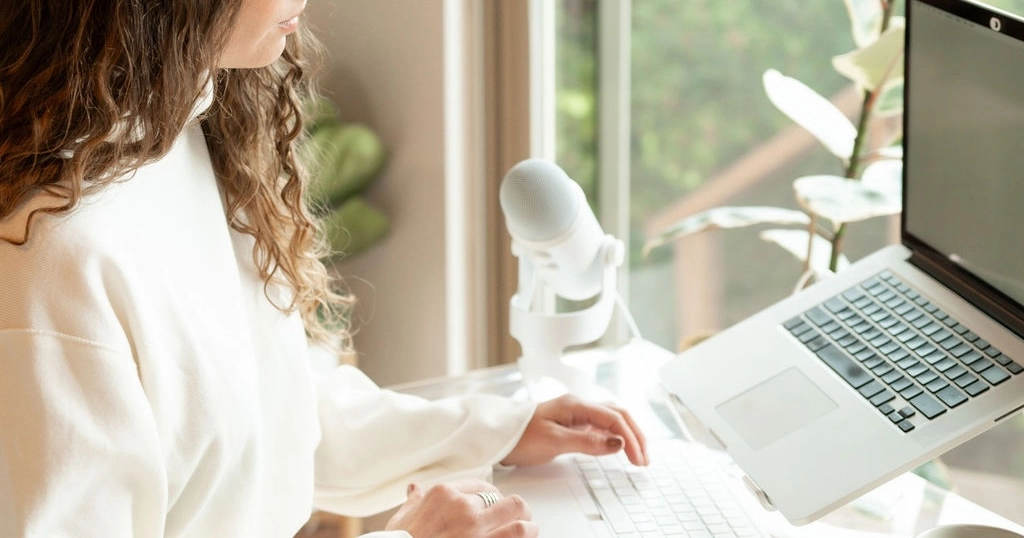 Woman with curly hair typing on a laptop keyboard next to a white microphone by a window with plants outside.