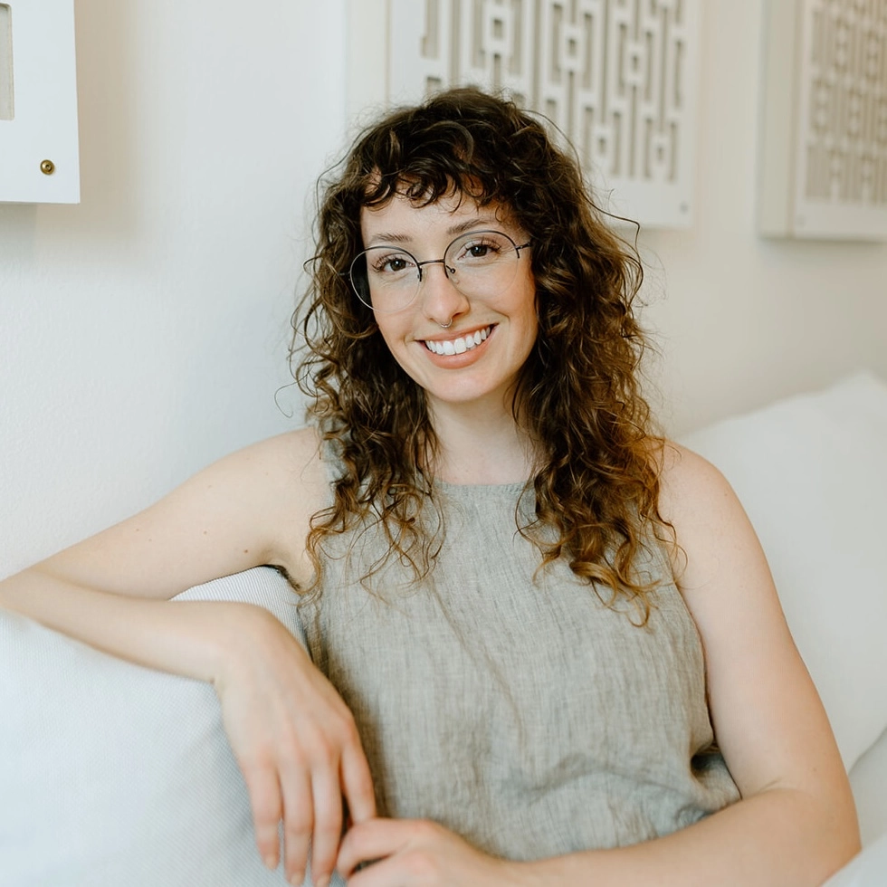 Smiling woman with curly brown hair, glasses, and a septum piercing sitting comfortably on a white couch.