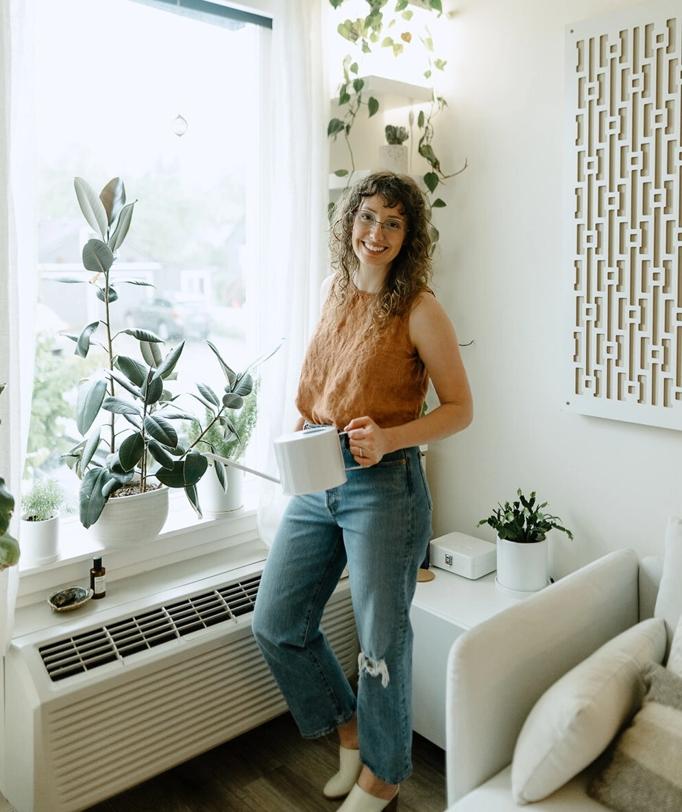 Woman with curly hair and glasses smiling while holding a white watering can near a window with indoor plants.