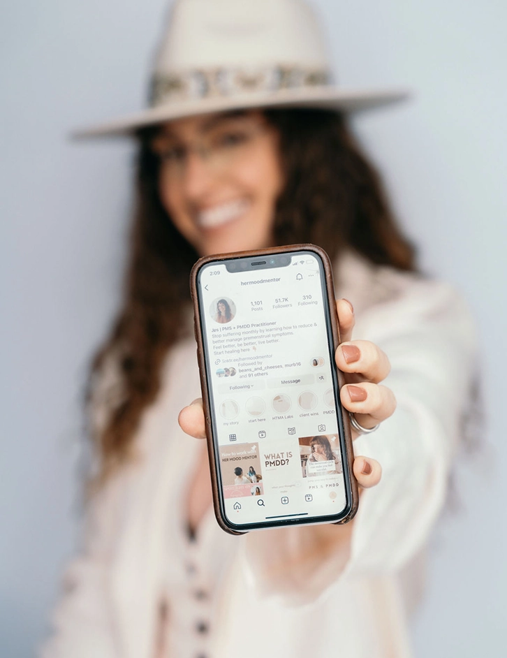 A woman wearing a hat and glasses holds a smartphone displaying the Instagram profile of 'hermoodmentor'.
