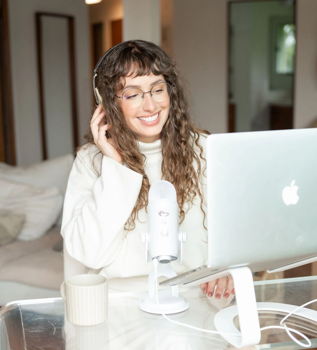 Smiling woman with curly hair and glasses wearing headphones, speaking into a white microphone while looking at a laptop on a glass table.