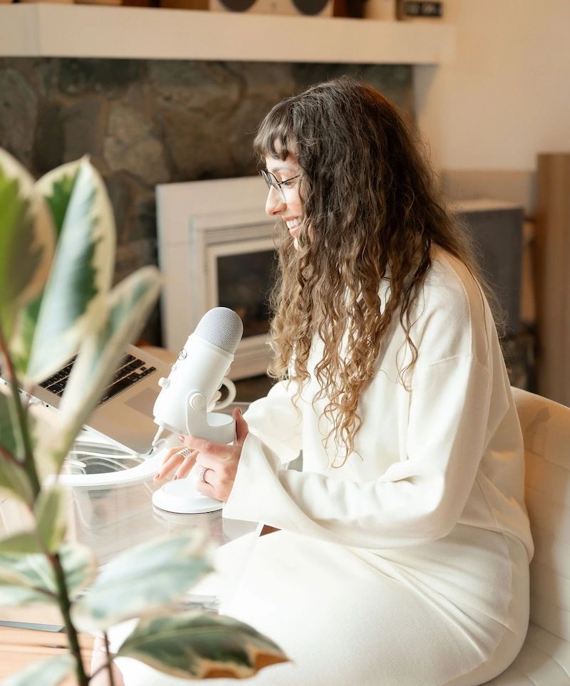 Woman with curly hair and glasses speaking into a white microphone in a cozy indoor setting with a laptop and plant nearby.