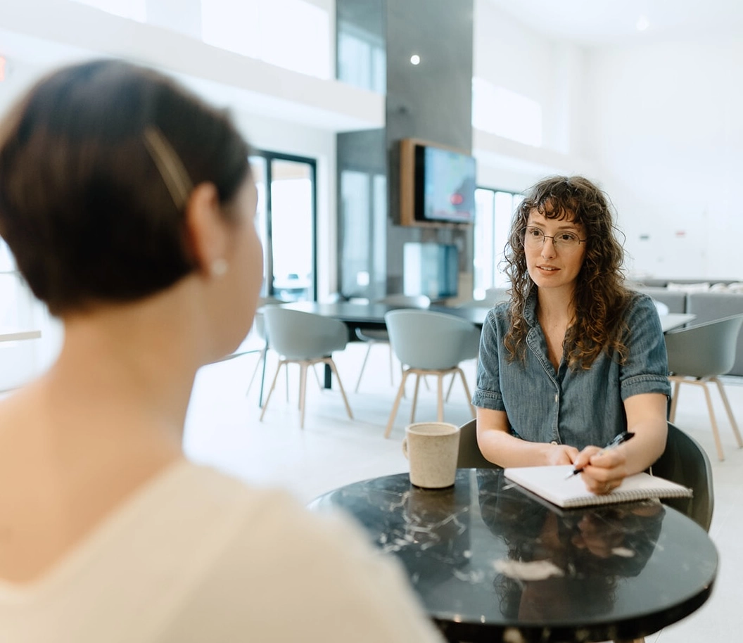 Two women sit at a round table in a modern office; one with curly hair and glasses takes notes while listening.