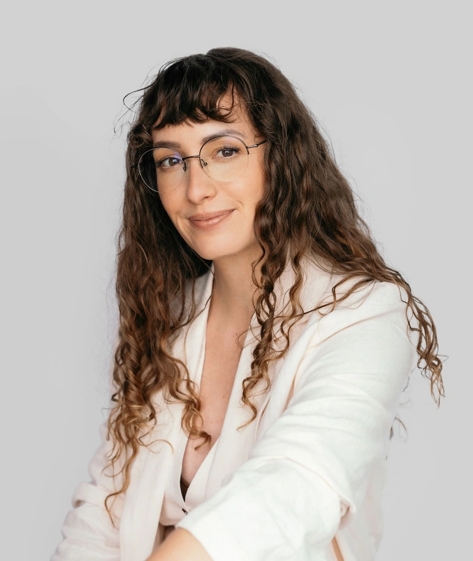 Smiling woman with long curly hair and glasses wearing a white blazer against a gray background.