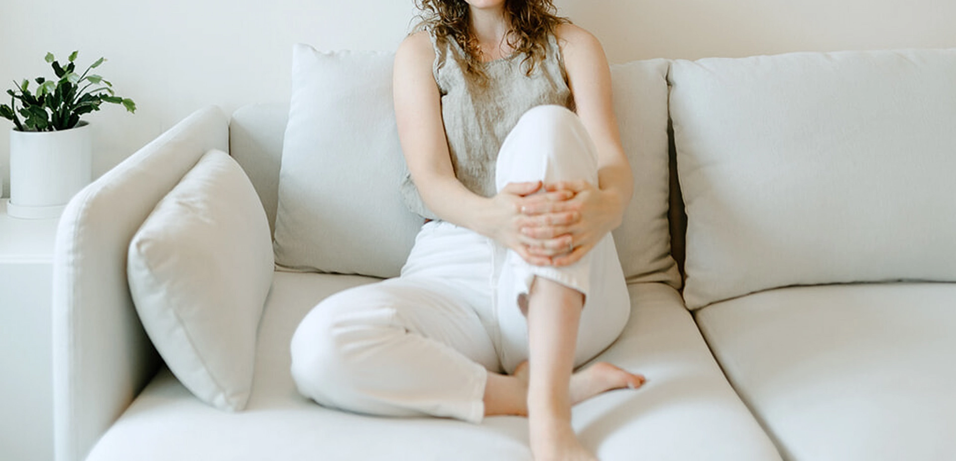 Smiling woman with curly hair and glasses sitting barefoot on a light gray sofa with folded hands around one knee.