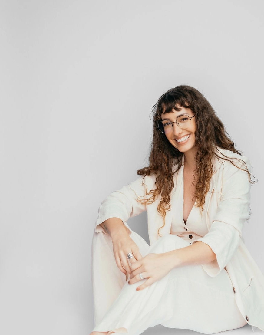Smiling woman with long curly hair and glasses sitting barefoot on the floor wearing a cream-colored suit.