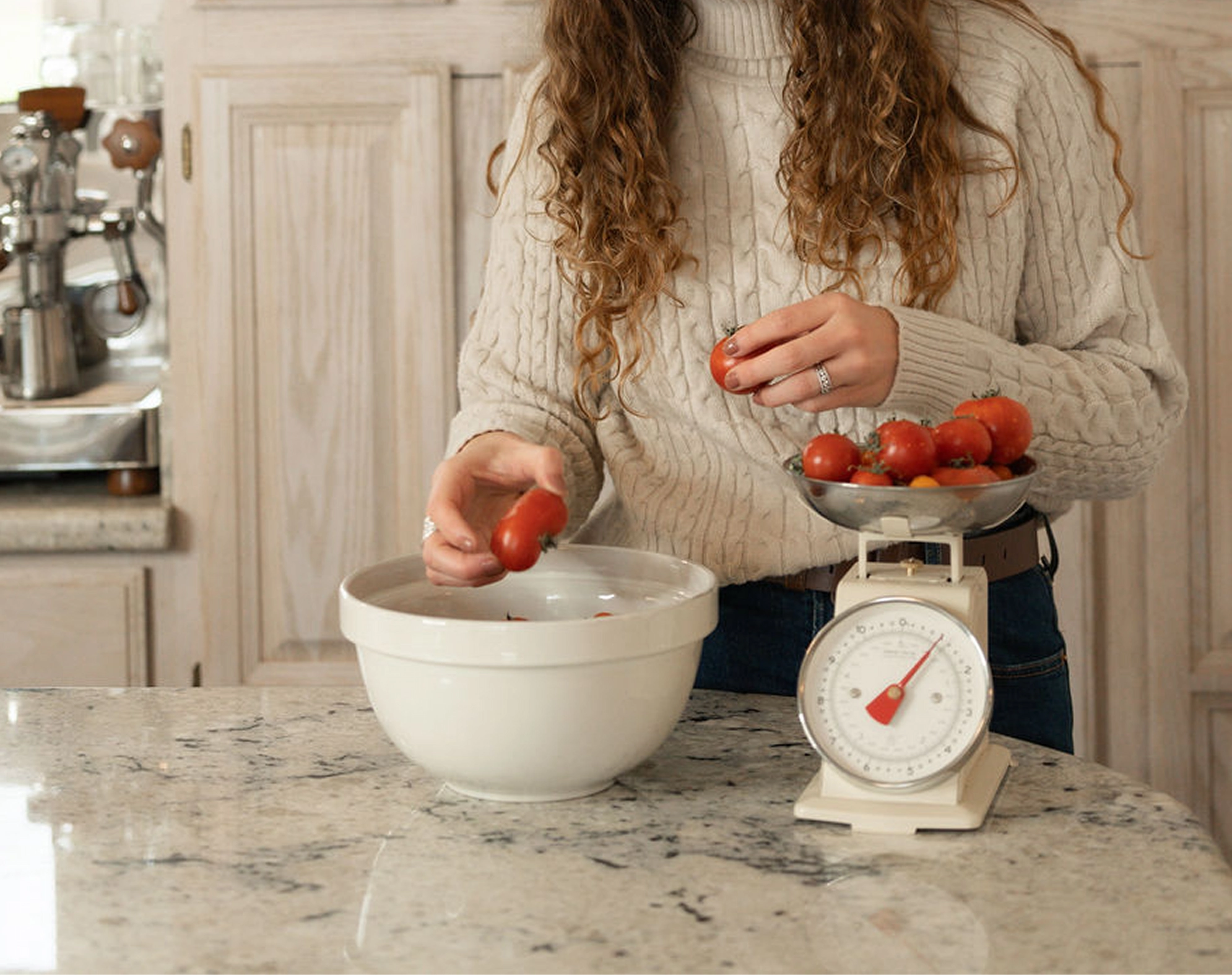 Smiling woman with curly hair and glasses holding cherry tomatoes next to a vintage kitchen scale in a rustic kitchen.