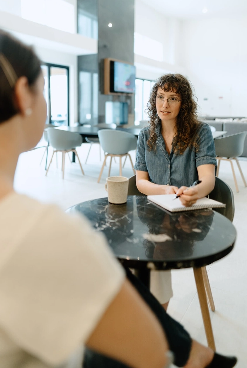 Woman with curly hair and glasses holding a pen and notebook while talking to another person at a round table in a bright modern room.