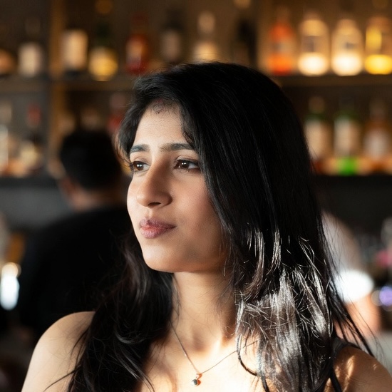 Portrait of a young woman with long dark hair looking thoughtfully to the side in a softly lit room with a blurred background of shelves with bottles.
