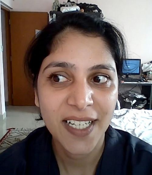 Close-up of a woman with dark hair looking to her left, smiling with visible teeth, in an indoor room with a computer and furniture in the background.
