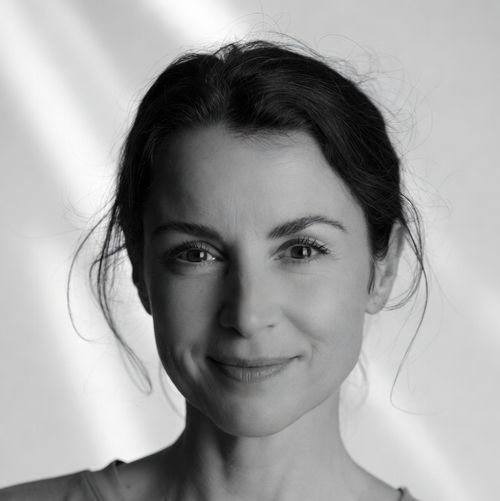 Close-up portrait of a woman with dark hair tied back, smiling softly with a blurred bright background.