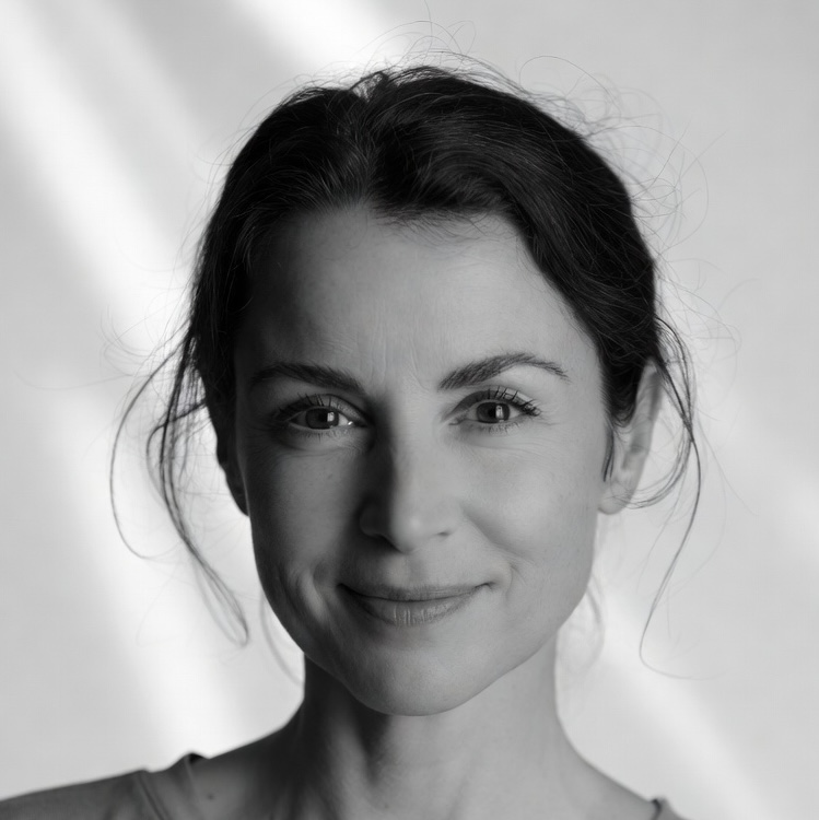 Close-up portrait of a woman with dark hair tied back, smiling softly with a blurred bright background.