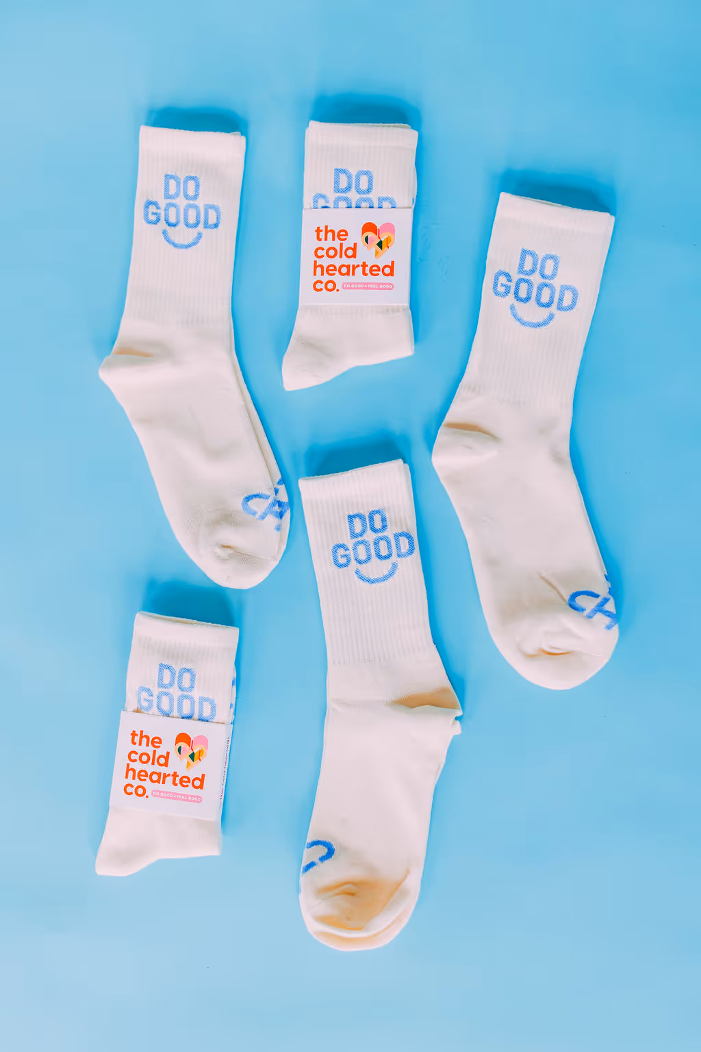 Five pairs of white socks with light blue 'DO GOOD' text and smiley faces, two pairs with 'the cold hearted co.' label on a blue background.