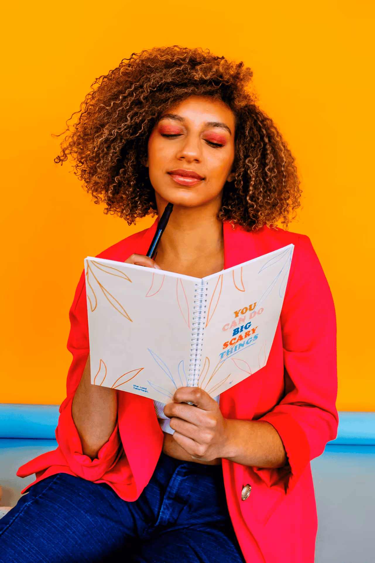 Woman with curly hair in a red blazer holding an open notebook that says 'You can do big scary things' against a yellow background.