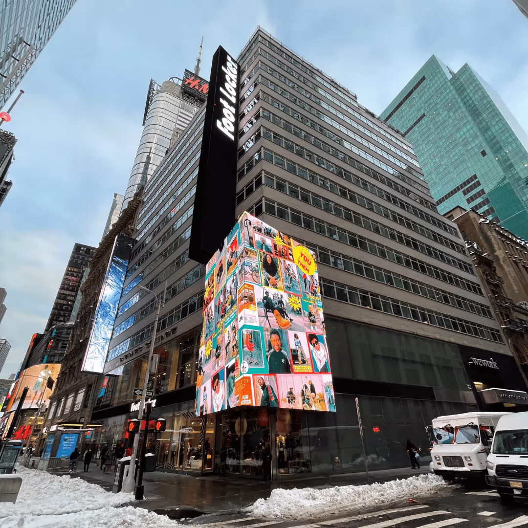 Foot Locker store on a snowy city street corner with large digital advertisements and tall buildings.