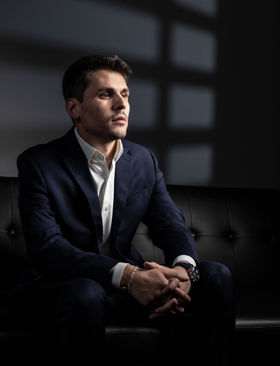 Charles W. Garcia in a navy blue suit sitting on a black leather sofa with hands clasped, looking contemplative.