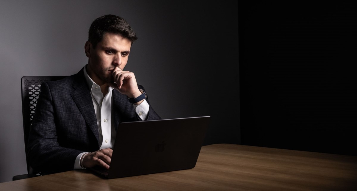 Charles W. Garcia in a dark blazer and white shirt sitting at a wooden table, focused on a laptop with a thoughtful expression in a dimly lit room.