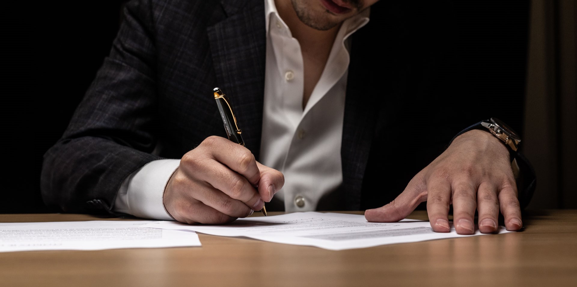 Charles W. Garcia in dark jacket and white shirt signing a document on a wooden table with a black and gold pen.