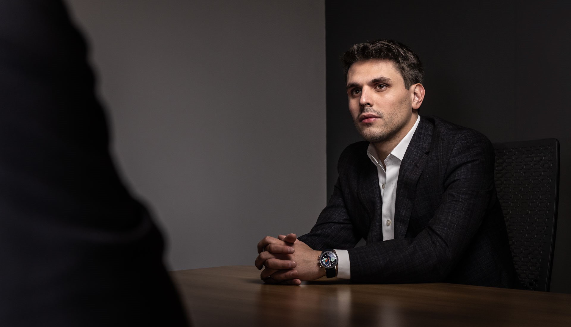 Charles W. Garcia in a dark checkered suit and white shirt sits at a wooden table with hands clasped, looking attentively.
