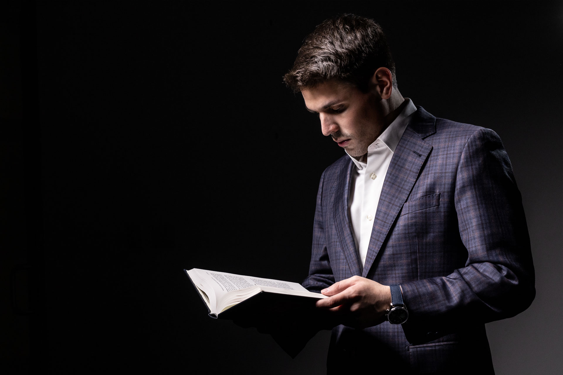 Charles W. Garcia in a dark checkered suit reading an open book against a dark background.