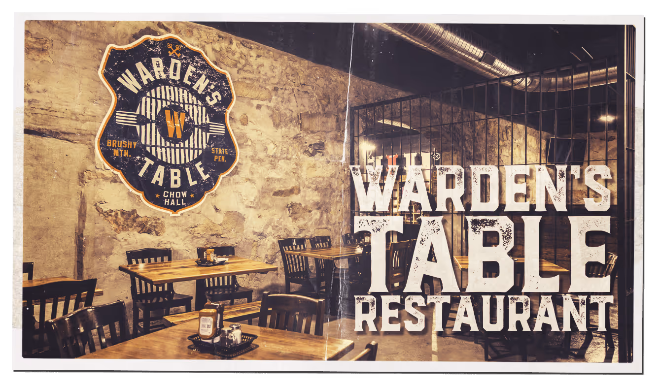 Interior view of Warden's Table Restaurant with rustic stone walls, wooden tables and chairs, and metal cage partitions and large text reading 'Warden's Table Restaurant'.