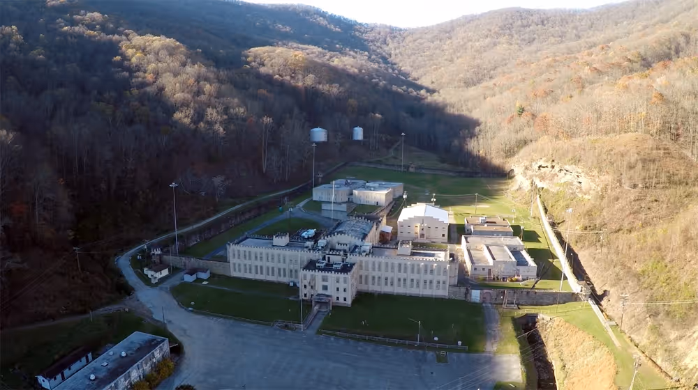 Aerial view of prison complex nestled in a valley surrounded by forested hills with some shadows cast on the buildings and landscape.