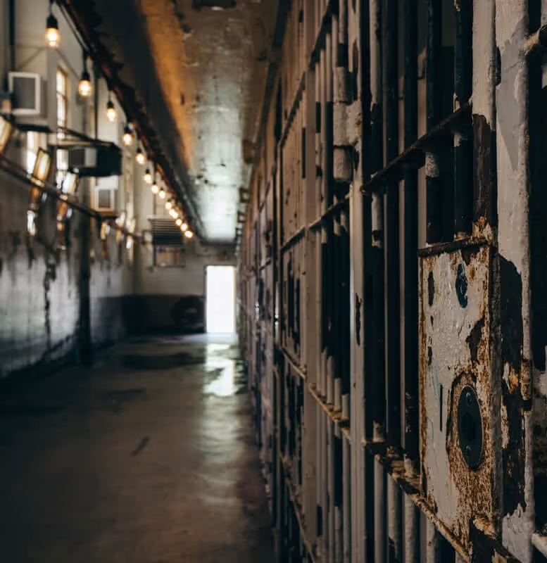 Dimly lit corridor in an old prison with rusted metal cell doors on the right and string lights along the ceiling on the left.