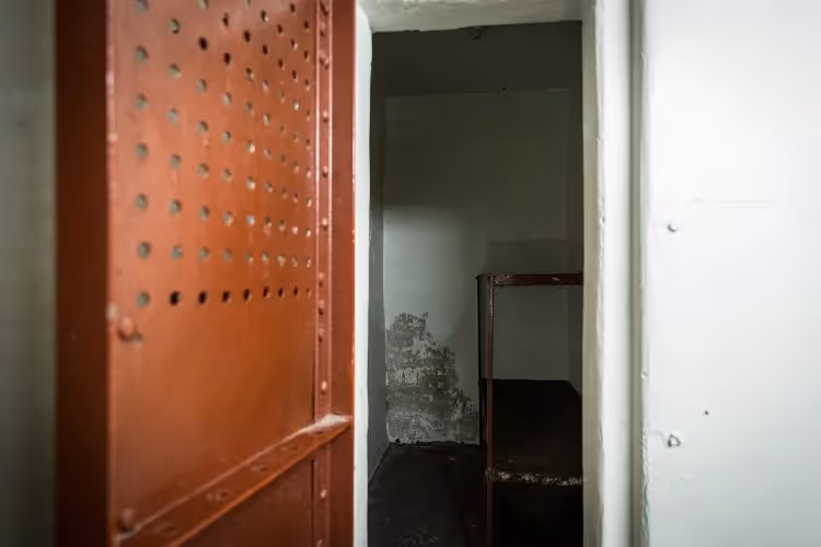 Interior of a small, empty prison cell with a rust-colored metal door and a single metal-framed bed.