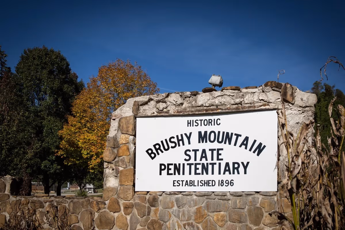 Stone monument with a white sign reading 'Historic Brushy Mountain State Penitentiary Established 1896' under a clear blue sky with trees in the background.