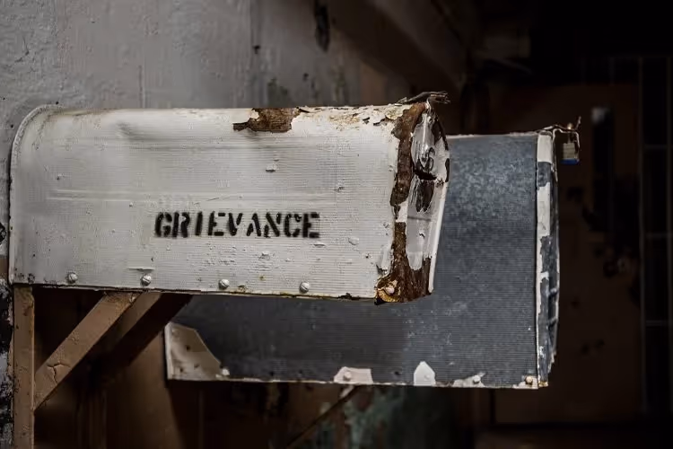 Rusty and peeling old metal mailbox labeled 'GRIEVANCE' mounted on a wall in a dimly lit, decayed setting.